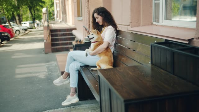 Lächelnde-Mädchen-mit-Smartphone-und-umarmung-Shiba-inu-Hund-im-Freien-im-Café