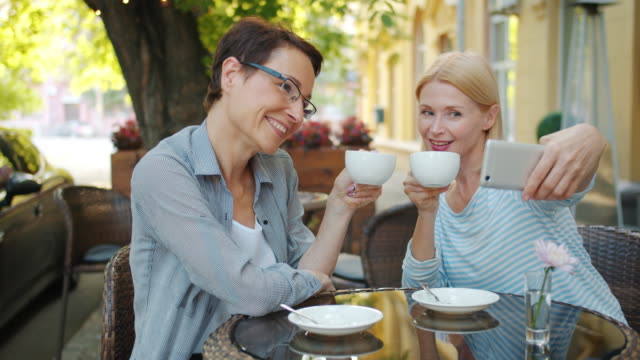 Reife-Damen-machen-Selfie-mit-Tassen-im-Open-Air-Café-halten-Kaffee-lächelnd
