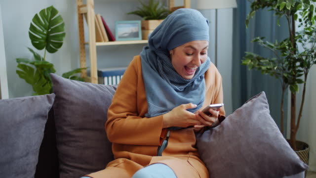 Fröhliche-Nah-Ost-Mädchen-in-Hijab-mit-Smartphone-lachen-Spaß-zu-Hause