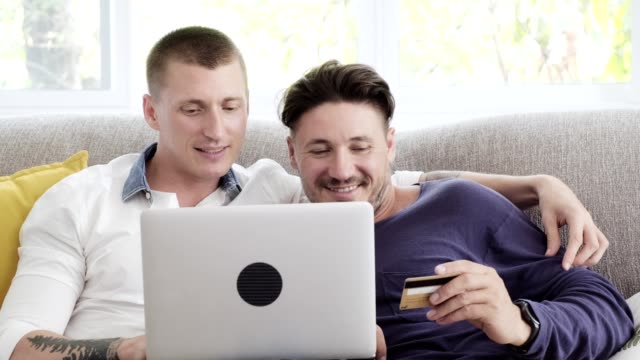 Gay-couple-relaxing-on-couch-using-laptop-computer.-Using-credit-card.