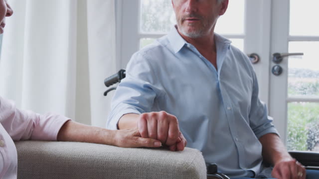 Close-Up-Of-Senior-Couple-With-Man-In-Wheelchair-Sitting-In-Lounge-At-Home-Talking-Together