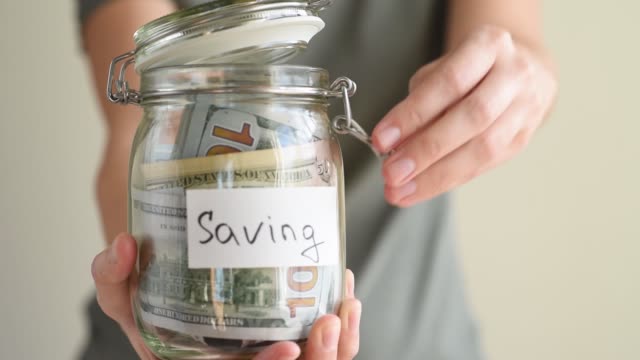 Woman-putting-paper-dollar-bill-in-a-glass-piggy-bank-with-inscription-Saving.-Save-money-concept