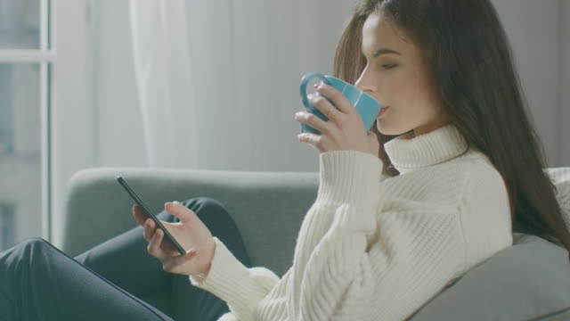 Beautiful-Young-Woman-Using-Smartphone,-Drinks-Tea-while-Sitting-on-the-Chair.-Sensual-Girl-Wearing-Sweater-Surfs-Internet,-Posts-On-Social-Media,-Shares-Pictures-while-Relaxing-in-Cozy-Apartment