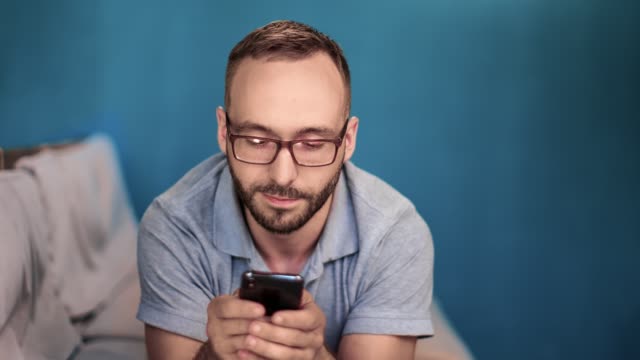 Smiling-man-in-glasses-lying-on-couch-chatting-use-smartphone.-Medium-close-up-shot-on-4k-RED-camera