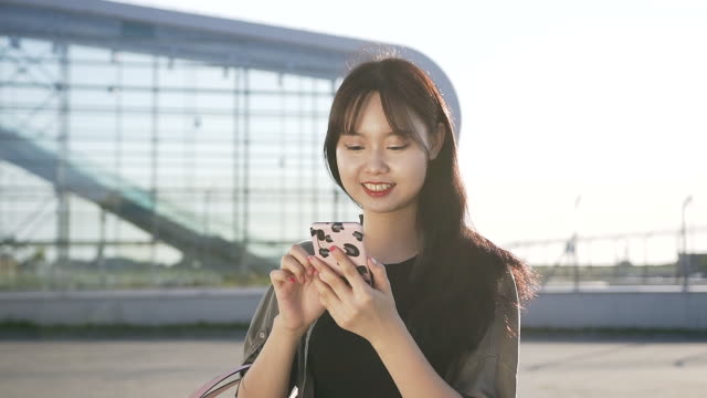 Hermosa-mujer-asiática-joven-sonriente-caminando-cerca-del-edificio-del-aeropuerto-y-usando-su-móvil