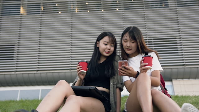 Atractivas-chicas-asiáticas-sonrientes-felices-sosteniendo-en-las-manos-café-y-mirando-el-teléfono,-sentado-en-la-hierba-cerca-de-un-gran-edificio-urbano
