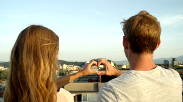 CLOSE-UP:-Couple-making-heart-shaped-symbol-and-kissing-on-rooftop-above-city
