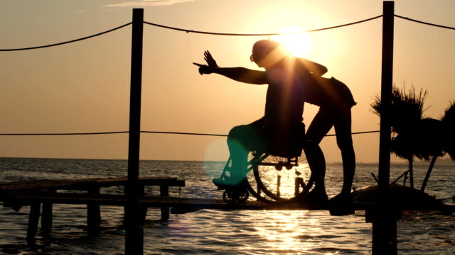 Fortunate-disabled-man-in-a-wheelchair-with-woman-looking-off-into-the-horizon-,background-of-beautiful-sundown-at-summer-travel