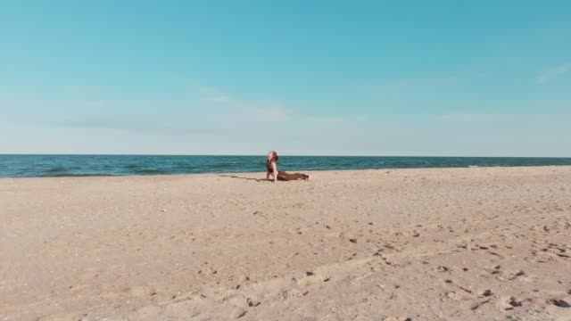 Drone-moves-behind-woman-in-sport-wear-holds-yoga-asana-position-on-the-sandy-sea-or-ocean-beach.-Windy-sunny-weather.-Aerial-view-of-peaceful-health-girl-performing-practice