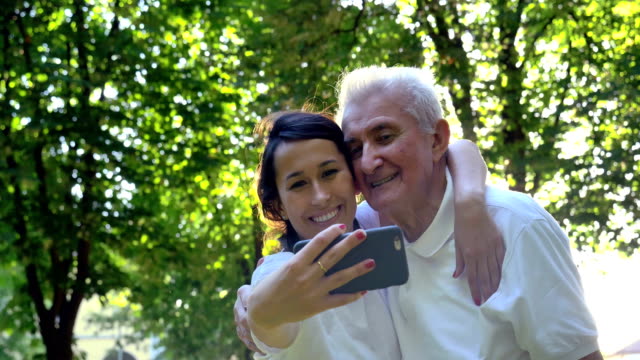 Una-niñera,-enfermera,-cuidar-de-los-ancianos,-una-chica-(mujer)-y-el-abuelo-se-sientan-en-un-libro-de-bolsillo-y-hacer-selfie,-en-el-parque.