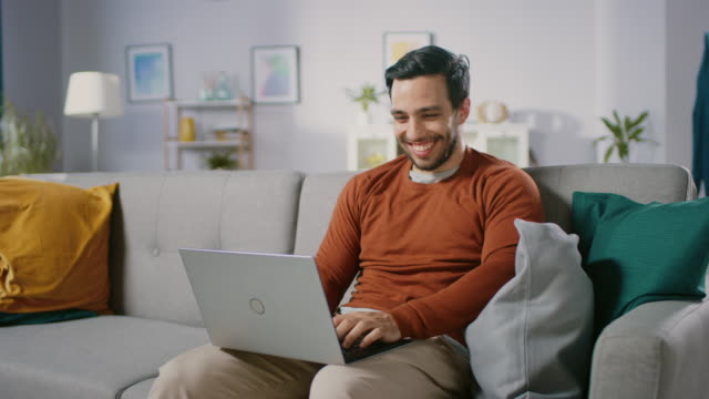 Fröhlichen-junger-Mann-zu-Hause-sitzen-auf-einem-Sofa-hält-Laptop-auf-dem-Schoß,-browst-durch-das-Internet,-soziale-Netzwerke,-macht-e-Shopping.