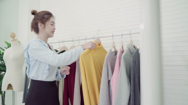 Asian-fashion-female-blogger-online-influencer-holding-shopping-bags-and-lots-of-clothes-on-clothes-rack-for-recording-new-fashion-video-broadcast-live-video-to-social-network-by-internet-at-home.