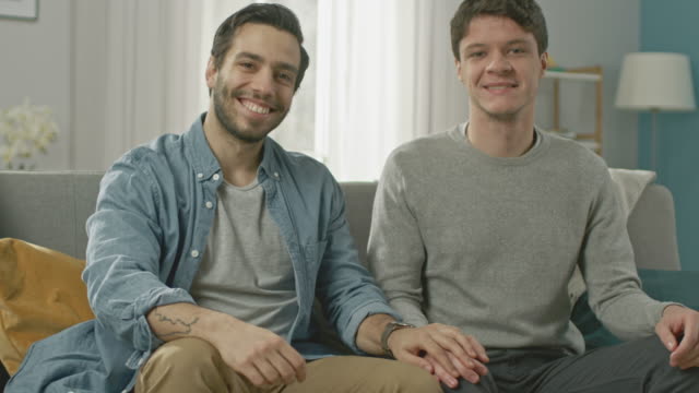 Cute-Attractive-Male-Gay-Couple-Sit-Together-on-a-Sofa-at-Home.-Boyfriend-Puts-His-Hand-on-Partner's.-They-are-Happy-and-Smiling.-They-are-Casually-Dressed-and-Their-Room-Has-Modern-Interior.
