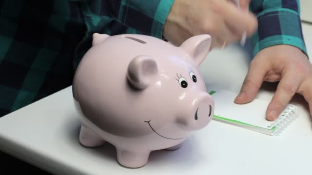 On-the-table-there-is-a-piggy-bank-in-the-form-of-a-pink-pig.-A-man-puts-a-coin-in-it,-makes-notes-in-a-notebook.