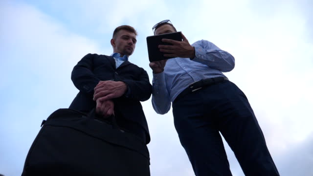 Young-businessman-showing-a-presentation-on-screen-of-tablet-pc-to-his-colleague-in-city.-Successful-entrepreneurs-working-on-digital-tablet-with-blue-sky-at-background.-Low-angle-view-Slow-motion