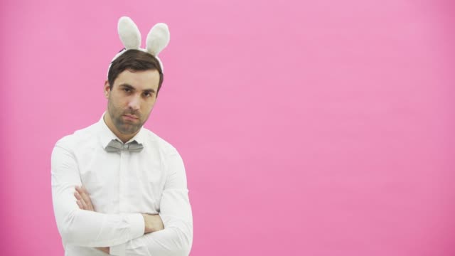 Young-handsome-man-standing-on-a-pink-background.-Dressed-in-a-white-shirt-with-rabbit-ears-on-his-head.-Putting-a-hand-on-the-hand,-serious,-looking-into-the-camera.