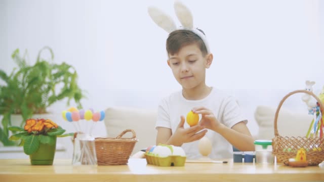 Lindo-niño-con-orejas-de-conejo-está-sosteniendo-huevos-de-Pascua-rosa-y-amarillo-imitando-como-los-ojos-y-luego-los-pone-de-nuevo-en-el-estante.