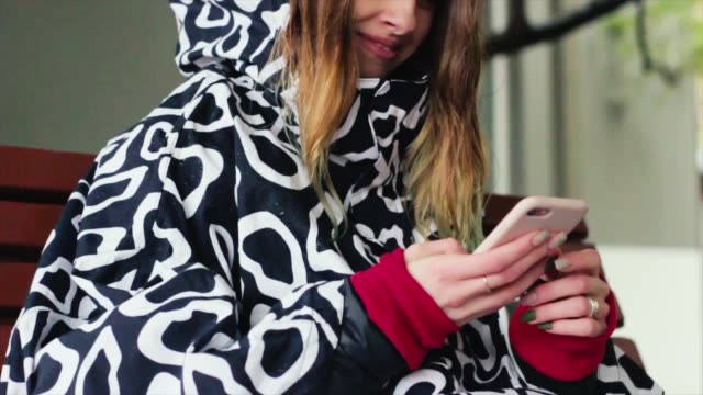 Lächelnde-Frau-im-Regenmantel-mit-Handy-und-Textnachrichten-in-sozialen-Netzwerken,-allein-sitzend
