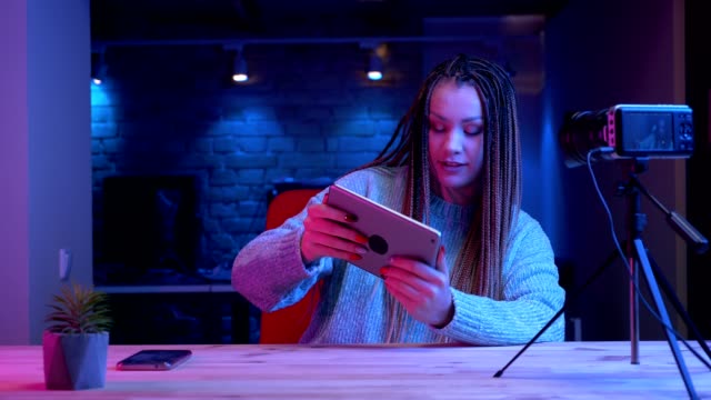 Closeup-shoot-of-young-attractive-female-blogger-with-dreadlocks-texting-on-the-tablet-streaming-live-with-the-neon-background-indoors