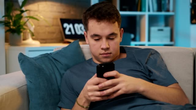 Un-guapo-adolescente-está-enviando-mensajes-de-texto-con-un-amigo-a-través-de-las-redes-sociales-en-su-teléfono-inteligente-mientras-está-sentado-en-un-sofá