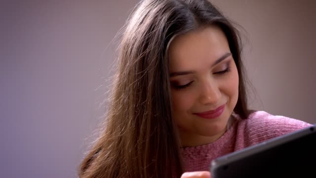 Closeup-shoot-of-young-pretty-brunette-caucasian-female-browsing-on-the-tablet-smiling-happily-and-looking-at-camera-in-a-cozy-apartment-indoors