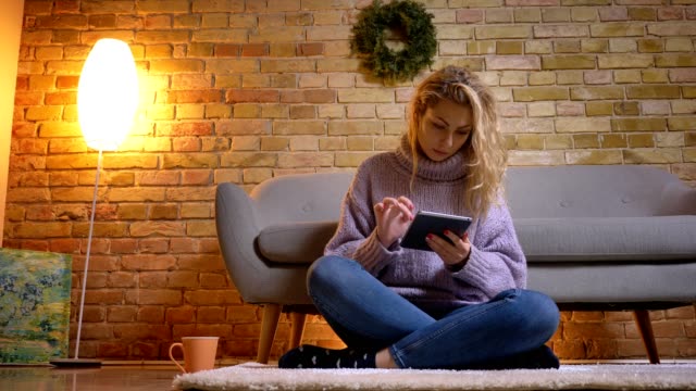 Closeup-top-shoot-of-adult-caucasian-blonde-female-using-the-tablet-while-sitting-on-the-floor-indoors-in-a-cozy-apartment
