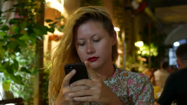 Pensive-girl-uses-a-smartphone,-writes-a-message,-chatting.-Woman-looking-for-information-in-the-phone-and-sad.-Sitting-in-restaurant-alone.-Evening-time,-lowlight.-Technology-and-entertainment-concept.-4k