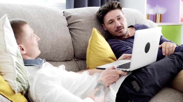Gay-couple-relaxing-on-couch-using-laptop-computer.-Talking.