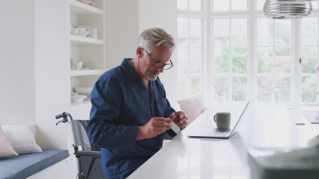Senior-Man-In-Wheelchair-At-Home-Looking-Up-Information-About-Medication-Online-Using-Laptop