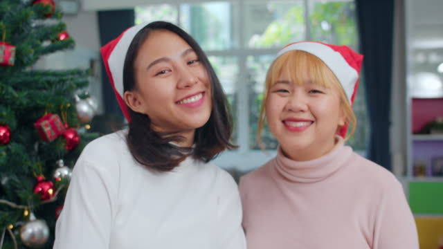 Asiatische-Lesbische-Paar-feiern-Weihnachtsfest.-LGBTQ-weibliche-teen-tragen-Weihnachten-Hut-entspannen-glücklich-lächelnd-Blick-auf-die-Kamera-genießen-Xmas-Winterurlaub-zusammen-im-Wohnzimmer-zu-Hause.