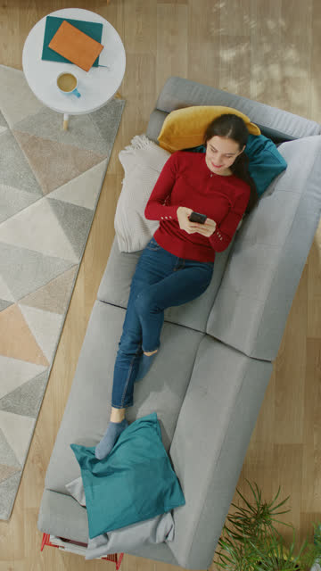 Junges-Mädchen-in-rotem-Pullover-und-blue-Jeans-liegt-auf-einem-Sofa,-mit-einem-Smartphone.-Sie-ist-glücklich-und-lächelt.-Top-Ansicht.-Vertikale-Bildschirmausrichtung-Video-9:16