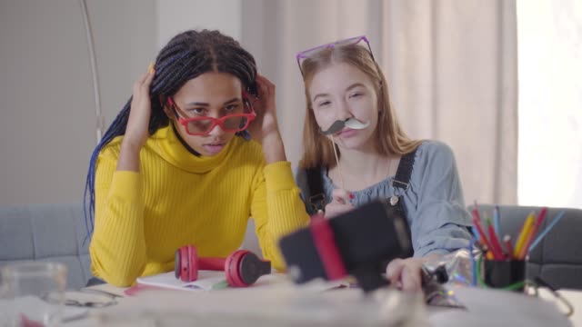 Afroamerikanische-Mädchen-in-roten-Brillen-und-ihr-kaukasischer-Freund-mit-falschen-Schnurrbart-sitzen-am-Tisch-und-schauen-auf-Smartphone-Bildschirm.-Teenager-machen-Selfie-Foto.-Spaß,-Glück,-Lebensstil.