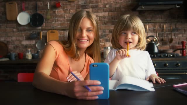 Mutter-und-Sohn-genießen-Online-Videoanruf-auf-dem-Smartphone,-während-Hausaufgaben-in-der-Schule-Copybook.-Sie-sitzen-in-der-Küche-am-Tisch.-Nahaufnahme