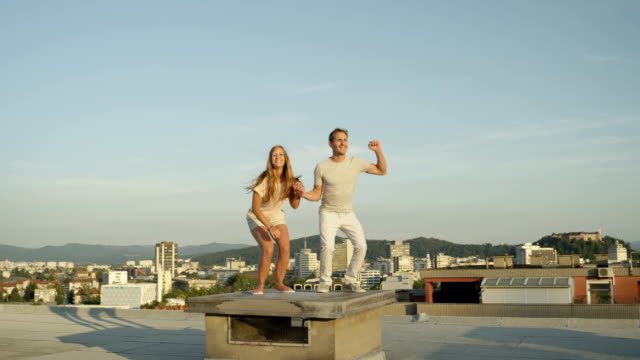 CLOSE-UP:-Girl-and-boy-holding-hands-and-jumping-on-rooftop-above-the-city