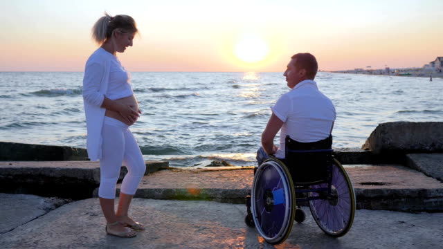 Spouse-in-wheelchair-holds-arm-woman-with-big-belly,-happy-couple-waiting-for-baby,-disabled-person-keeps-hand-of-wife