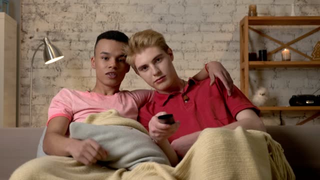 Two-gay-guys-are-sitting-on-the-couch-and-watching-TV,-a-Caucasian-guy-wants-to-switch-the-channel-but-an-African-guy-stops-him.-LGBT-lovers,-a-multinational-couple,-a-happy-gay-family,-a-home-cosiness-concept.-Look-at-the-camera-60-fps