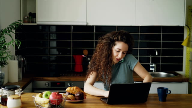 Young-curly-pretty-positive-caucasian-woman-working-on-laptop-and-talking-on-phone-sitting-in-modern-kitchen,-she-is-printing-and-talking-in-one-and-the-same-time