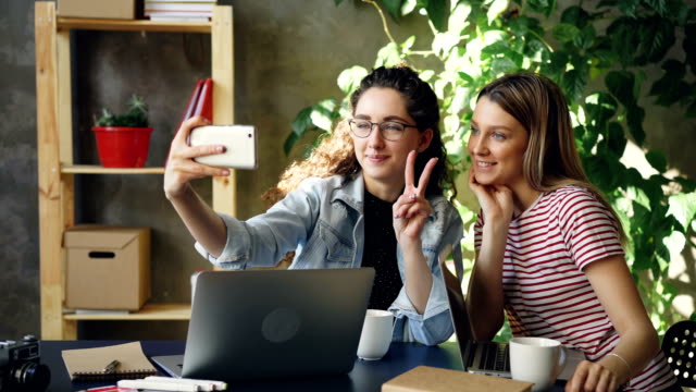 Junge-Unternehmer-sprechen-und-machen-Selfie-mit-Smartphone-beim-Sitzen-am-Schreibtisch-im-modernen-Büro.-Sie-posiert-und-lachen,-dann-Fotos-ansehen.