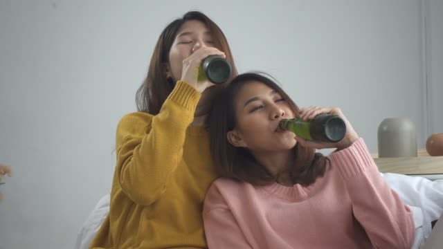 Glücklich-asiatische-Lesben-LGBT-Ehepaar-am-Sofa-sitzen-und-Biertrinken-im-Wohnzimmer.-Frauen-feiern-Sie-mit-ihrer-Freundin-zu-Hause.