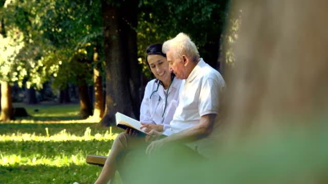 Una-niñera,-enfermera,-cuidar-a-los-ancianos-una-chica-(mujer)-y-el-abuelo-se-sientan-en-un-libro-y-leer-un-libro,-en-el-parque.
