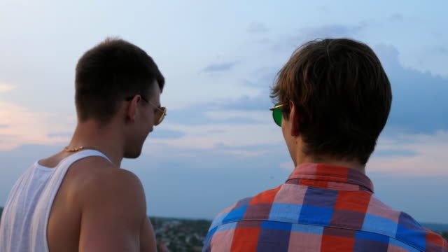 Dolly-shot-of-young-male-couple-in-sunglasses-standing-on-the-edge-of-rooftop-and-talking.-Handsome-gay-boys-rest-on-roof-of-high-rise-building-and-enjoying-beautiful-cityscape.-Close-up-Slow-motion