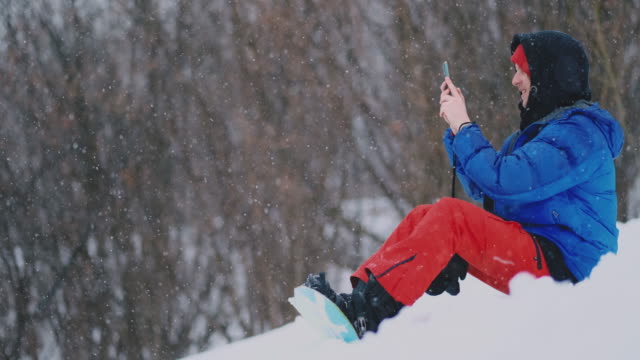 Männliche-Snowboarder-sitzen-auf-dem-Schnee-und-machen-Fotos-am-Telefon-schöne-Landschaft-des-Resorts-für-soziale-Netzwerke