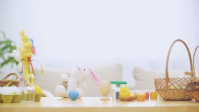 Little-boy-is-hiding-under-the-wooden-table,-full-of-Easter-decorations:-basket,-yellow-chicken,-colorful-eggs-pains-and-paint-brush.-Boy-is-playing-with-a-cute,-soft-white-bunny-with-pink-ears,-on-the-table.-Bunny-is-holding-a-paint-brush.