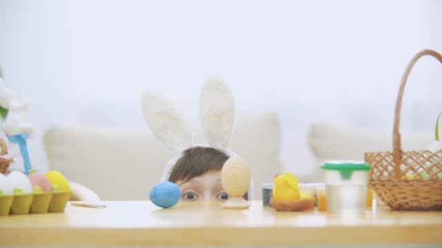 Little-playing-boy-w-is-hiding-under-the-wooden-table,-full-of-Easter-decorations.-Boy-is-winking-and-smiling-with-his-eyes,-hiding-his-face.
