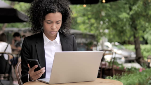 Afrikanische-Frau-mit-Smartphone-und-Laptop-in-Outdoor-Cafe