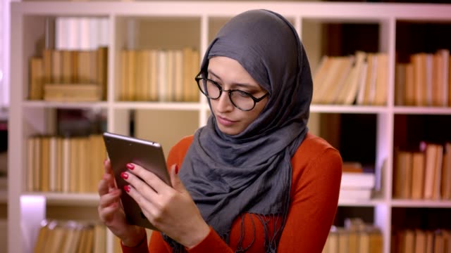 Sesión-de-primer-plano-de-joven-atractiva-estudiante-musulmana-en-hiyab-mecanografía-en-la-tableta-de-pie-en-el-interior-de-la-biblioteca