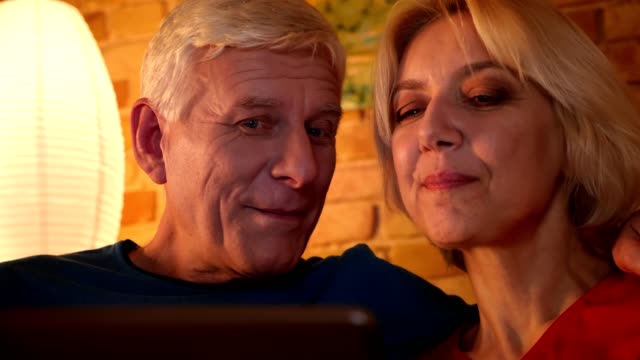 Closeup-shoot-of-aged-happy-couple-browsing-the-network-on-the-tablet-sitting-on-the-sofa-resting-together-indoors-in-a-cozy-flat