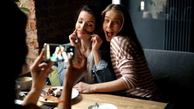 Happy-young-ladies-having-fun-in-cafe-taking-photos-with-smartphone-posing