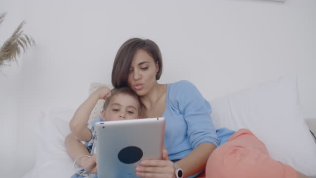 Im-weißen-Schlafzimmer-schauen-Mama-und-Sohn-auf-den-Tablet-Bildschirm-und-lachen.-Glückliche-Familie-im-Bett-am-Morgen-lesen-ein-Buch-und-klicken-Sie-auf-das-Touchpad-des-Computers
