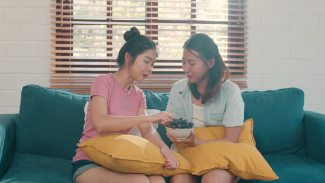 Pareja-lesbiana-asiática-comer-comida-saludable-mientras-está-acostado-sofá-en-la-sala-de-estar-en-casa.